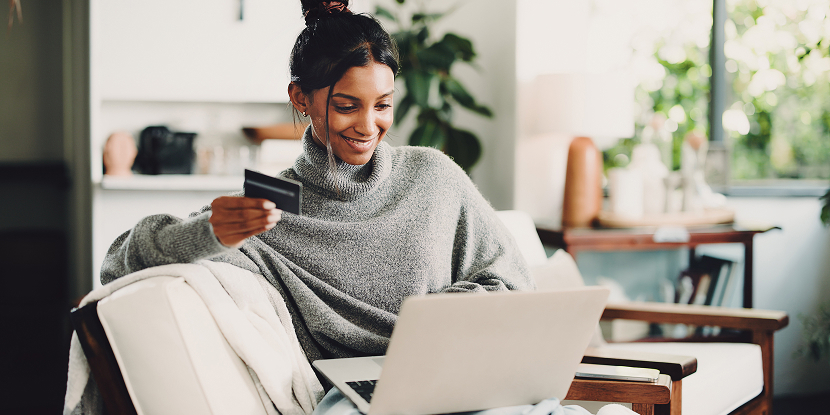 Cliente souriante tenant une carte bancaire devant son ordinateur portable, assise dans un fauteuil